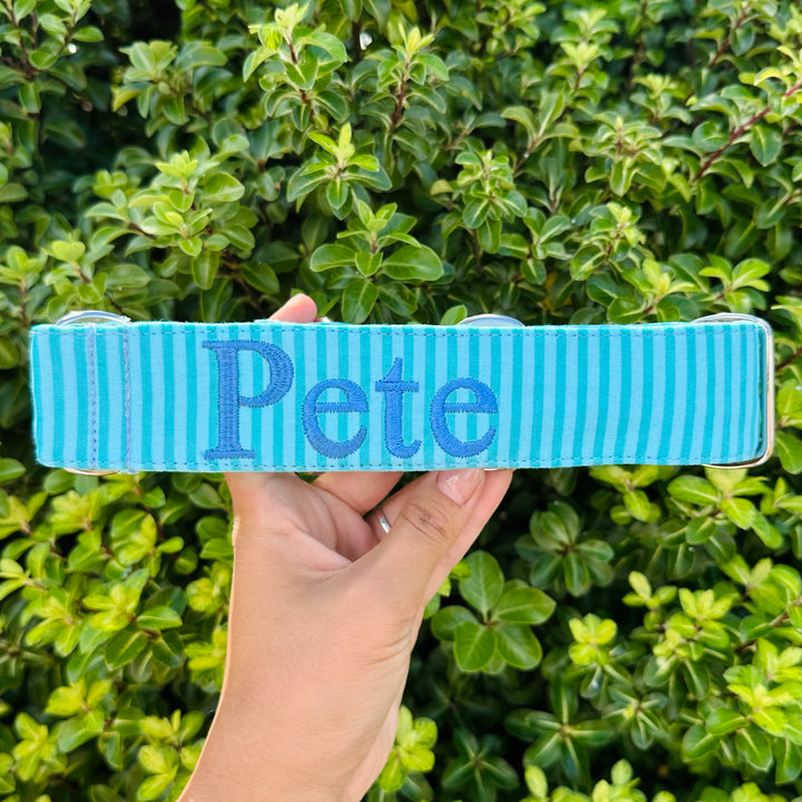 A person holding a custom-made martingale dog collar in light blue with teal horizontal stripes, embroidered with the name 'Pete' in bold blue letters. The background features a lush green hedge.