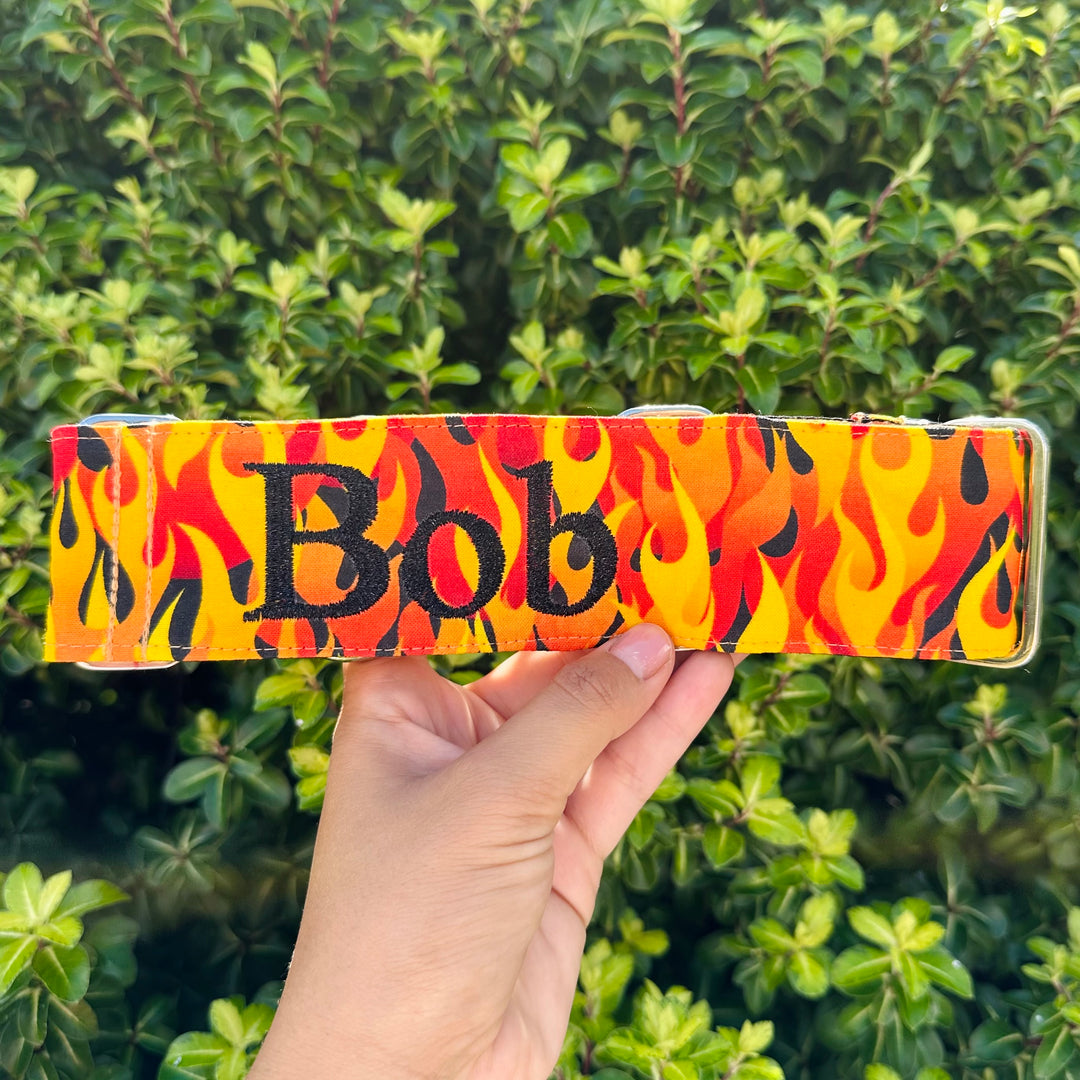 A person holding a custom-made martingale dog collar with a vivid flame pattern in red, orange, and yellow. The name 'Bob' is prominently displayed in bold, black letters against the fiery background. The setting includes a backdrop of lush green hedge.