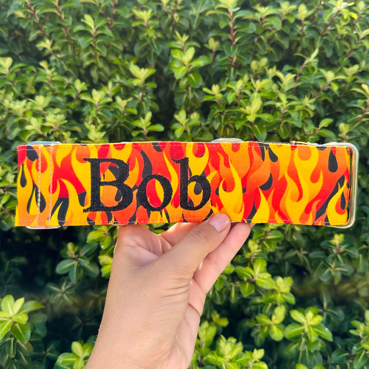 A person holding a custom-made martingale dog collar with a vivid flame pattern in red, orange, and yellow. The name 'Bob' is prominently displayed in bold, black letters against the fiery background. The setting includes a backdrop of lush green hedge.